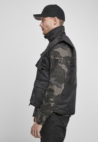 Svart ermeløs vest med camo-mønstrede lange ermer, med høy krage, framlommer og knappedetaljer. Brukt med en svart caps.