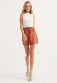 Witte geribbelde mouwloze top met een oranje corduroy minirok met knopen aan de voorzijde, afgebeeld tegen een neutrale achtergrond.