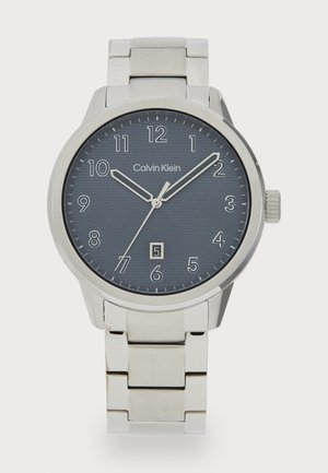 Montre en acier inoxydable de couleur argent de Calvin Klein avec un cadran rond bleu marine, des index horaires blancs et un affichage de date.