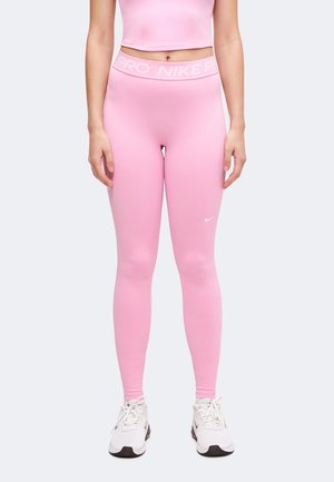 Kobieta w różowych legginsach Nike Pro z wysokim stanem i białych butach Nike, stoi z rękami swobodnie opuszczonymi wzdłuż ciała na jednolitym tle.
