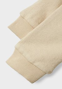 Beige gebreide trin gemaakt van zacht materiaal met een ribbelpatroon aan de mouwen. De stof voelt zacht en lichtgewicht aan, met een gladde afwerking.