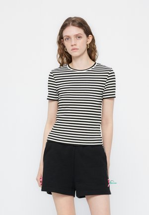 Jeune femme aux cheveux ondulés portant des boucles d'oreilles créoles en or, un haut à manches courtes rayé noir et blanc, et un short noir avec des rubans colorés.