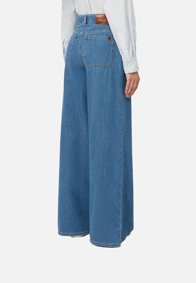 Persona che indossa jeans blu a vita alta e gamba larga con tasche posteriori e una camicia a righe chiara con maniche lunghe, in piedi di spalle.