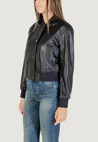 Giubbotto bomber in pelle nera con bottoni a pressione, polsini a costine e tasca anteriore; abbinato a jeans in denim blu. Texture liscia e design aderente.