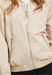 Frau trägt eine beige Reißverschlussjacke mit goldenen Reißverschlussdetails über einem weißen Hemd und schwarzen Hosen, gezeigt von der Mitte des Oberkörpers abwärts.