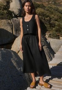 Un crop top en lin noir avec des boutons sur le devant et une jupe noire, ample, à taille élastique ; porté avec des sabots à bout ouvert beiges sur un chemin en bois.