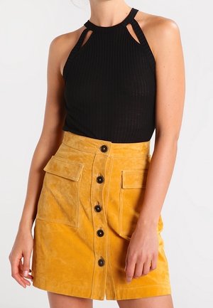 Vrouw draagt een mouwloos zwart topje met uitsparingen in sleutelgatstijl bij de hals en een geel hoogtaille rok met knopen aan de voorkant en grote voorzakken.