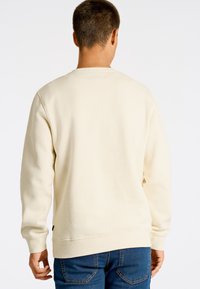 Sudadera color crema de manga larga y cuello redondo, con puños y dobladillo acanalados, llevada con jeans azules. Vista posterior.