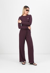 Bordeaux-ribbet jumpsuit med lange ermer og rund hals, med justerbar snor i midjen. Brukt med svarte flip-flops.