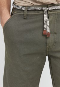 Pantalons vert olive avec une surface texturée, présentant une ceinture à motifs blancs et noirs et un accent en cuir près de la taille.