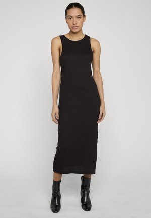 Femme debout, portant une robe midi noire côtelée sans manches avec un col rond et des bottines à talons noires, sur un fond uni.