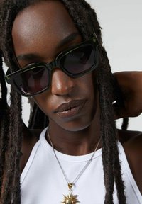 Zwarte rechthoekige zonnebril met getinte lenzen en heldergroene accenten, gedragen door een persoon met lang, donker haar, in een witte top en gouden zonnependant.