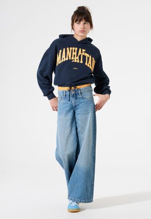Jonge vrouw in een marineblauwe "Manhattan" hoodie, met wijde lichtblauwe jeans en pastelblauwe sneakers, staand tegen een witte achtergrond.