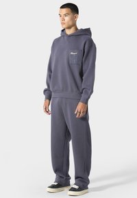Sudadera morada con bolsillo frontal y detalles gráficos, combinada con pantalones de chándal holgados a juego. La tela parece suave y texturizada. Zapatillas negras.
