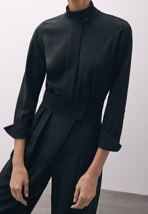 Woman wearing a black long-sleeve jumpsuit with a high collar, button closure, belt detail, and folded cuffs, standing against a plain background.