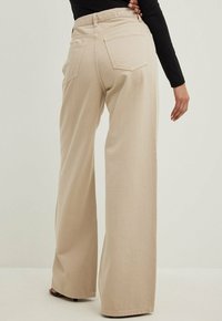Pantaloni beige a gamba larga con vita alta, dotati di tasche posteriori e un tessuto liscio e texturizzato. Accentuati da un dettaglio con logo in vita.