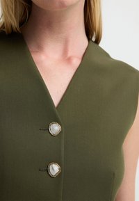 Oliwkowozielony bezrękawnik z głębokim dekoltem w kształcie litery V oraz dwoma beżowymi guzikami ze złotymi akcentami przy dekolcie. Gładka faktura.