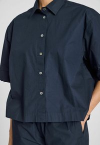 Camicetta blu navy a maniche corte con bottoni, realizzata in cotone liscio, con vestibilità comoda e classica chiusura con bottoni, dotata di tasche laterali.