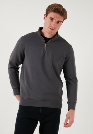 Siva zip-up dukserica s visokim ovratnikom, dugim rukavima, rebrastim manžetama i glatkom teksturom, nosi se preko crne majice.