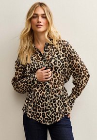Camisa con botones de estampado de leopardo en tela ligera; cuenta con mangas largas, cintura con cinturón y dobladillo recto. La paleta de colores incluye tonos marrones y negros.