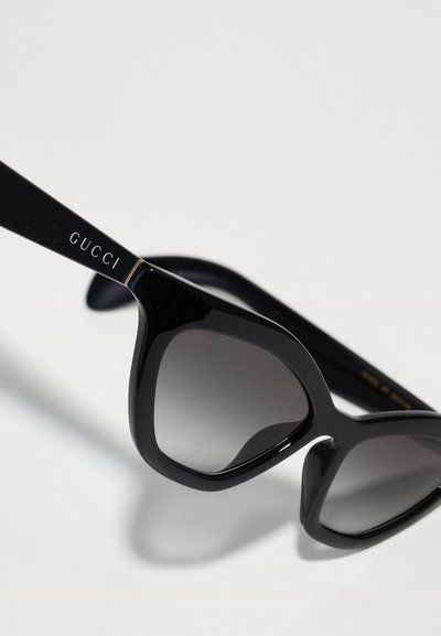 Occhiali da sole neri a forma di occhio di gatto, con lenti sfumate, montatura in plastica lucida e sottili dettagli dorati sulle tempie, firmati "Gucci."