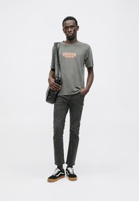 Grijze katoenen t-shirt met de tekst "STRANGER THINGS", zwarte schoudertas met straps, donkergrijze broek en zwarte sneakers met witte accenten.