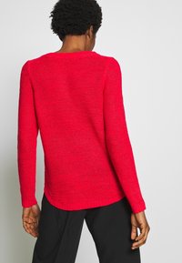 Pull rouge en maille avec manches longues, encolure ronde et finition texturée ; présente des poignets et un ourlet côtelés, avec une silhouette décontractée.