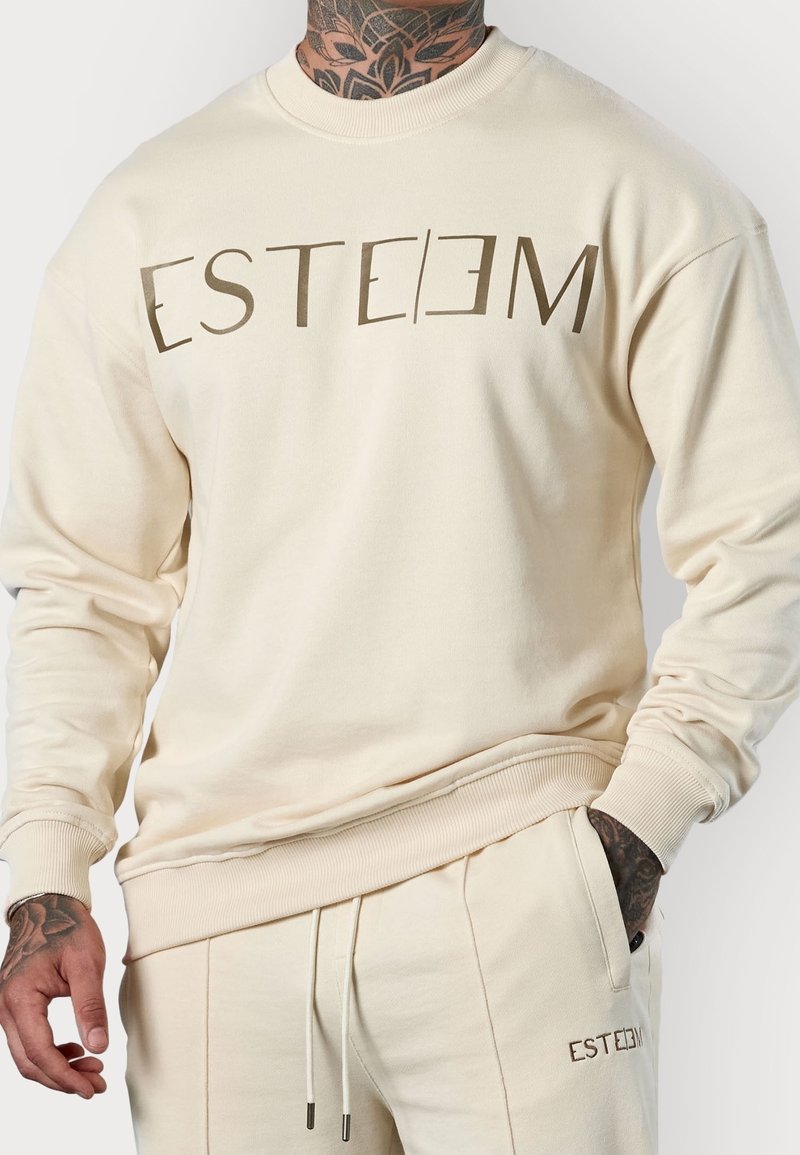 Sweat-shirt beige et pantalon assorti avec le texte "ESTEEM", porté par une personne tatouée, main dans la poche, sur fond blanc.