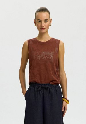 Femme aux cheveux tirés en arrière portant un haut marron sans manches à motifs, un pantalon bleu marine avec cordon, des boucles d’oreilles anneaux dorés et des bracelets.