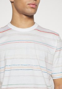 Młody mężczyzna ubrany w biały T-shirt w cienkie, wielokolorowe poziome paski, pokazany od szyi do połowy torsu na jednolitym tle.