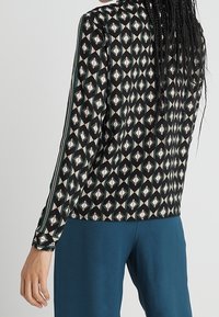 Maglia a maniche lunghe in tessuto nero con un motivo geometrico in tonalità di bianco, rosso e verde. Presenta strisce laterali a contrasto.