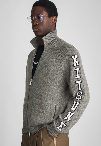 Gestrickter grauer Reißverschluss-Pullover mit hohem Kragen, versehen mit weißem "KITSUNE"-Text auf dem Ärmel und einer strukturierten Oberfläche. Zwei Fronttaschen.