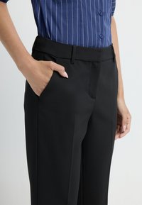 Zwarte tailorpantalons met een gladde textuur, voorzien van een zijzak, vlakke voorkant en een sleek silhouet. Gecombineerd met een blauw gestreept shirt.