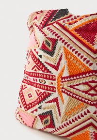 Štvorcový dekoratívny vankúš s vyšívanými geometrickými vzormi. Dominantné farby sú červená, oranžová, čierna a béžová. Textúrovaná látka.