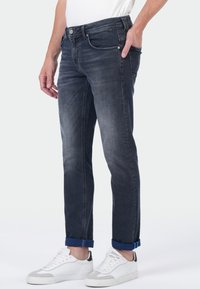 Man in donkere gewassen jeans met omslagen en witte sneakers, staand met een hand in zijn zak tegen een effen witte achtergrond.