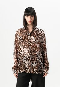 Mulher com cabelo preto liso, vestindo uma camisa oversized de estampa de leopardo e calças pretas, de pé contra um fundo branco liso.