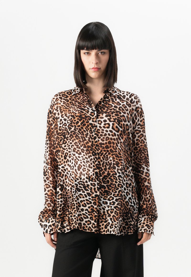 Mulher com cabelo preto liso, vestindo uma camisa oversized de estampa de leopardo e calças pretas, de pé contra um fundo branco liso.