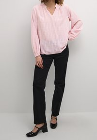 Blouse rose clair à manches longues avec col en V et détail froncé, associée à un pantalon droit noir et des chaussures à talons carrés brillantes noires.