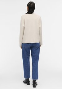 Beige genser med høy hals, kombinert med blå tapered jeans og svarte ankelstøvler, sett bakfra. Teksturen er myk og glatt.