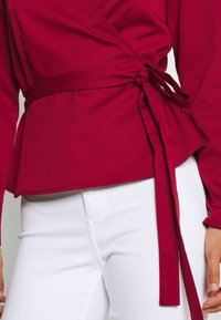 Haut portefeuille bordeaux à manches longues avec fermeture à nouer. Le tissu a une texture lisse, avec un bas à volant et un éclat subtil.