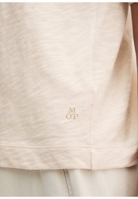 Ourlet de chemise en coton beige avec de petites lettres "M OP" discrètes centrées juste au-dessus de la couture cousue sur un pantalon beige clair.