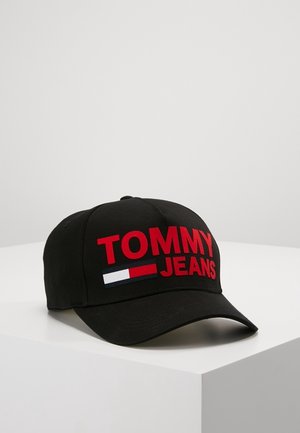 Casquette de baseball noire avec le texte "TOMMY JEANS" en rouge et un logo rectangulaire rouge, blanc et bleu à l'avant, posée sur une surface blanche.