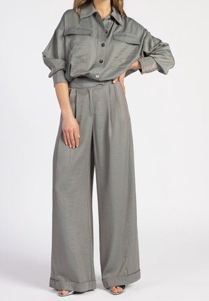 Nainen, joka pukeutunut löysään, harmaaseen nappipaitaan, jossa on rintataskut, ja vastaaviin leveälahkeisiin housuihin, seisoo yhdellä kädellä lanteillaan.