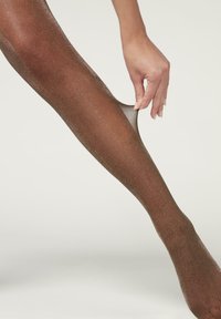 Bruna transparenta strumpbyxor med en glittrig finish, som visar en slät textur och en hand som drar i midjebandet för att justera passformen.