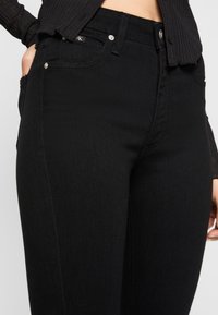 Person i svarta högmidjade jeans med synliga framfickor och en kort svart ribbad tröja, händerna i fickorna.