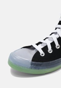 Zapatilla de lona negra con cordones blancos, tapa de puntera de goma transparente y suela verde translúcida. Presenta ojales plateados y texturas contrastantes.
