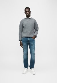 Γκρι πλεκτό πουλόβερ με ρίγες και ραγκλάν μανίκια, συνδυασμένο με μπλε τζιν κανονικής γραμμής και λευκά αθλητικά παπούτσια. Το μοντέλο στέκεται με τα χέρια στις τσέπες.