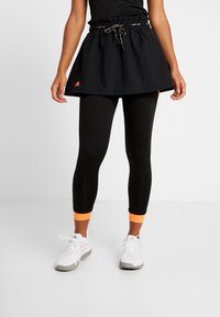 Person i svart sportkjol över svarta leggings med orange ankelmanschetter och vita sportskor, står på en vanlig vit golv.
