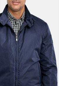 Chaqueta azul marino con textura suave y brillante y cierre de cremallera en la parte delantera. Presenta un cuello amplio y se lleva sobre una camisa a cuadros.