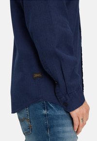 Close-up of a person wearing a navy blue long-sleeve shirt with a "Made for Daily Adventures" label, and blue denim jeans.